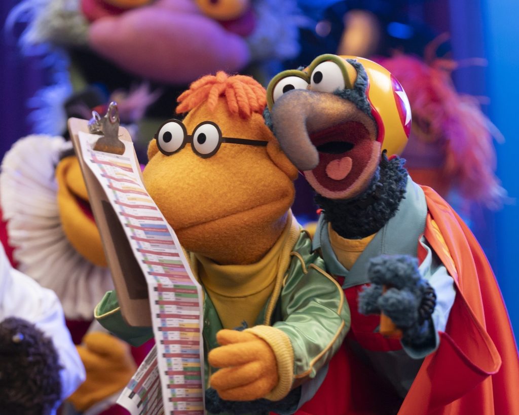 Scooter, wearing a yellow turtleneck and a green jacket, and Gonzo, wearing a helmet and cape, look at a clipboard of future bits for the show