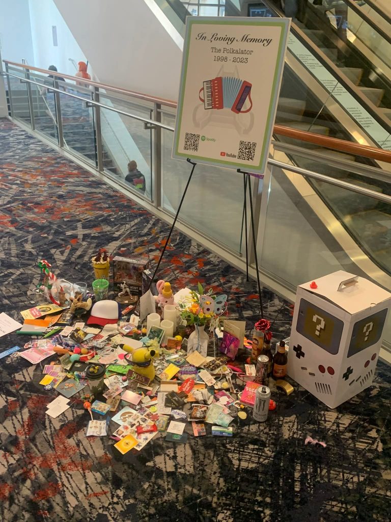 A cardboard sign with an accordion that reads "In Loving Memory" surrounded by small gifts. 