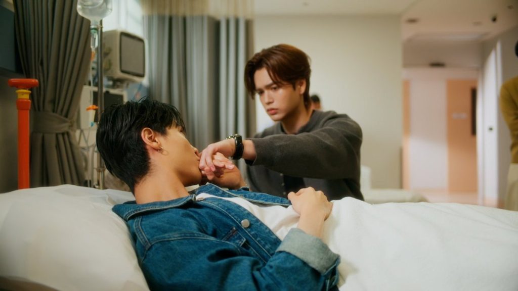 Tim is in the hospital after slipping and hitting his head. Pai sits, pouting, at his beside. Tim is kissing the back of Pai's hand.