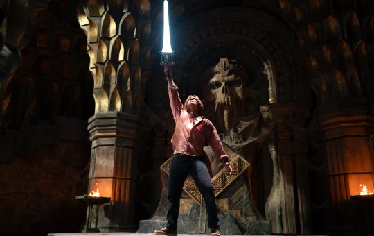 a young white man, wearing a pink shirt and black pants, stands in front of an alter with a skull in it, sword held high